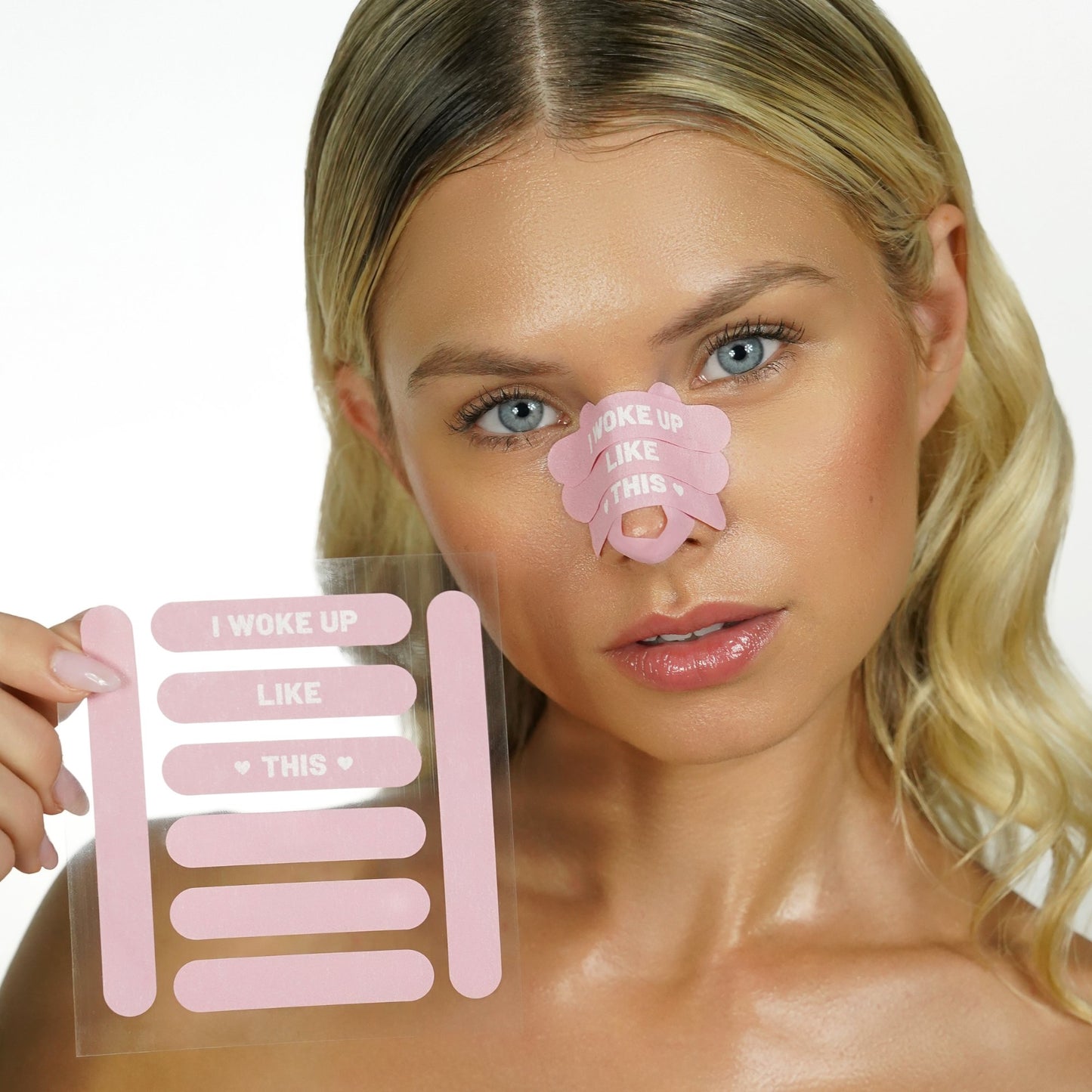 Woman holding a sheet of pink 'recovery babe' adhesive pre-cut nose strips with 'I woke up like this' text, with matching strips on her nose, on a white background