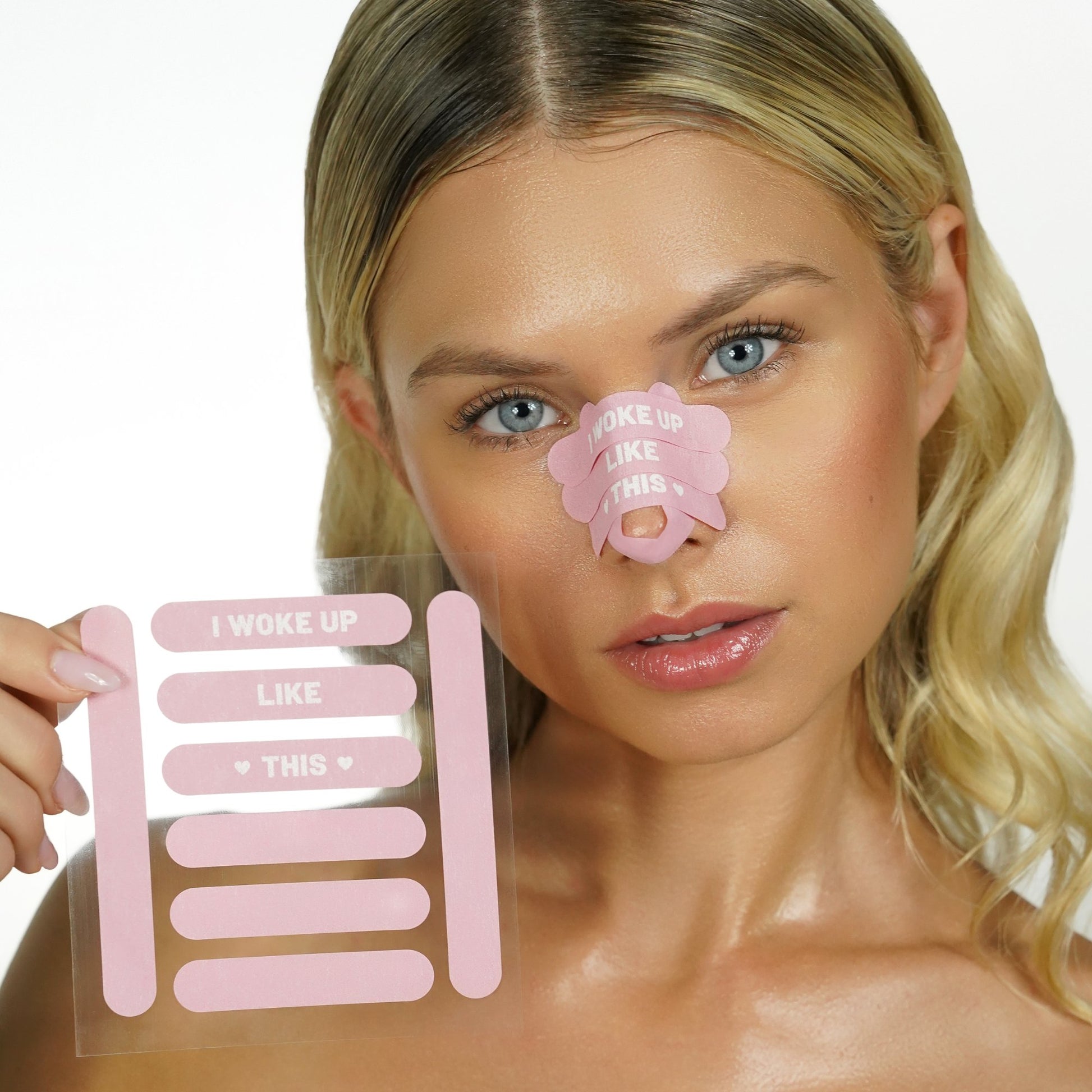 Woman holding a sheet of pink 'recovery babe' adhesive pre-cut nose strips with 'I woke up like this' text, with matching strips on her nose, on a white background